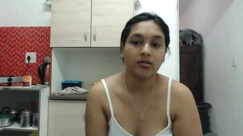 Media: Video of a young woman with dark hair, medium skin tone, wearing a white spaghetti-strap top and a necklace, standing in a kitchen with light wooden cabinets, red patterned wall, and various kitchen items visible.