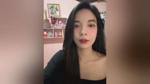 Media: Video of a young East Asian woman with long black hair, light skin, and red lipstick, wearing a black top. Background includes pink walls, shelves with plush toys, and a framed picture.