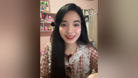 Media: Video of a smiling young Asian woman with long black hair and fair skin, wearing a checkered blouse and lace collar, standing in a pink room with shelves of toys and a stuffed animal.