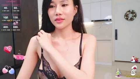 Media: A video of an Asian woman with straight black hair, wearing a black lace bra, standing in a modern kitchen with white cabinets, hearts, and a Christmas tree in the background.