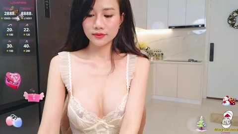 Media: Video of an East Asian woman with long black hair, wearing a revealing lace bra, standing in a modern kitchen with white cabinets, smiling.