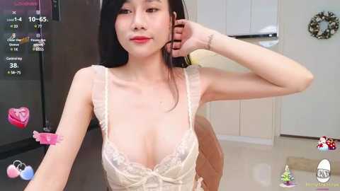 Media: Video of an East Asian woman with fair skin, long black hair, and red lipstick, wearing a sheer white lace lingerie bra, sitting in a modern kitchen.