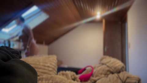 Media: Video of a cozy, dimly lit attic room with wooden ceiling panels, soft beige walls, and a pile of blankets and pillows. A person, partially visible, is wearing a pink shoe and black clothing.