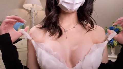 Media: Video of a woman in a white lace bra, face covered by a white mask, holding straps, in a softly lit bedroom with a lamp, floral arrangement, and dresser.