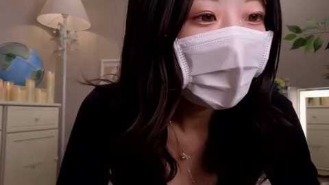 Media: Video of an East Asian woman with long black hair, wearing a white medical mask, black top, and a silver necklace, standing indoors with a beige lamp, a white dresser, and a globe in the background.