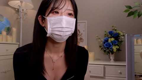 Media: Video of an East Asian woman with straight black hair and a white surgical mask, wearing a black top in a room with beige walls, white furniture, and a blue floral arrangement.