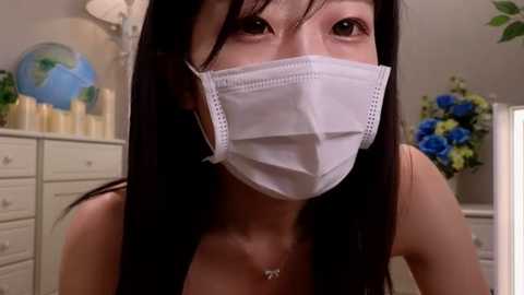 Media: Video of an Asian woman with long black hair, wearing a white surgical mask, in a brightly lit room with white furniture, blue flowers, and candles.