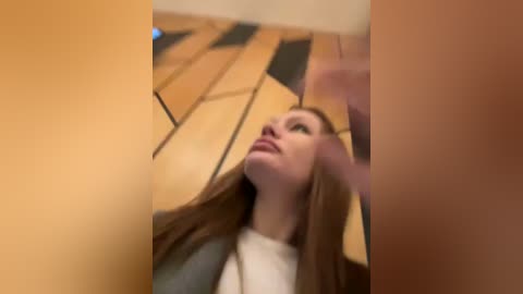 Media: Video of a young woman with long brown hair, wearing a white top, leaning against a wooden wall with geometric patterns. The image is slightly blurred and taken from an angle, capturing a casual, indoor setting.