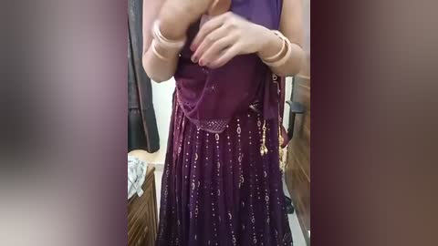 Media: Video of a light-skinned woman in a purple, sequined sari, with gold jewelry, standing in a room with beige walls and a wooden dresser.