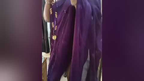 Media: A video of two people in purple sarees, with one person holding a phone and a medal, standing indoors. The background shows a beige wall and a wicker basket.