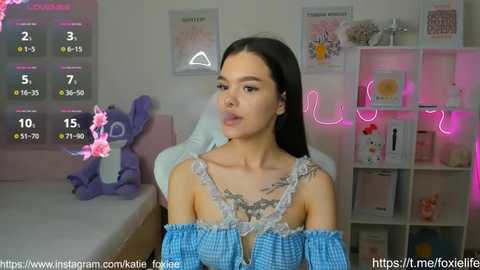 Media: A video of a young woman with long black hair, wearing a blue off-shoulder dress, sitting in a pink-lit room with a bed, plush toys, and a shelf in the background.