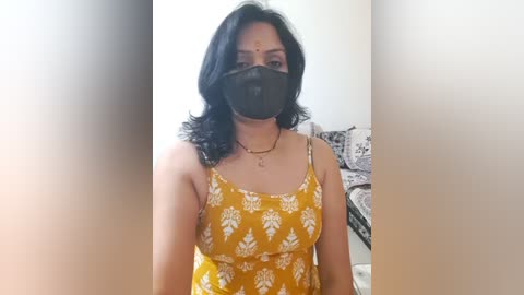 Media: Video of a woman with medium skin tone, black hair, wearing a yellow floral-patterned dress, black mask, and a bindi, standing in a home with a white wall and patterned sofa in the background.