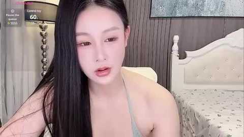 Media: Video of an East Asian woman with long, straight black hair, wearing a light gray camisole, seated on a bed with white, tufted headboard in a softly lit bedroom.