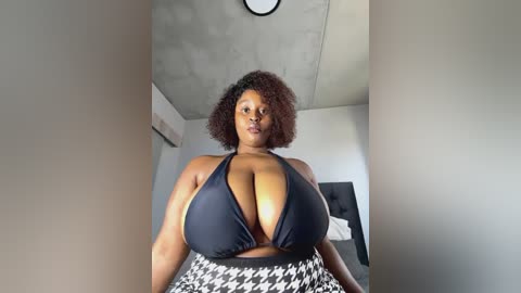 Media: Video of a plus-sized Black woman with natural hair, wearing a black halter top and houndstooth skirt, standing in a minimalist room with exposed concrete ceiling and light-colored walls.