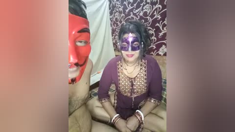 Media: Video of a woman in a purple saree, adorned with a purple mask and gold bling, sitting cross-legged on a beige bed with floral curtains. A red-faced man with a black beard and moustache is partially visible on the left.