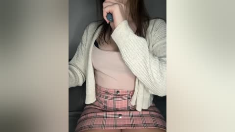 Media: Video of a woman with light skin, wearing a beige cardigan over a pink tank top, and a pink plaid skirt, holding a blue device to her face, in a dimly lit room.