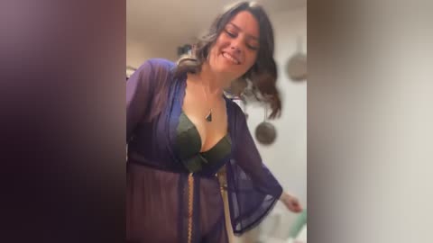 Media: Video of a woman with wavy brown hair, wearing a sheer blue robe over a green lace bra, smiling slightly. Background shows a blurry kitchen with white walls.