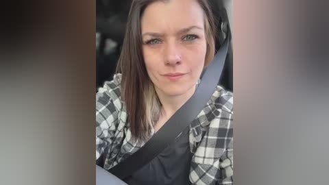 Video of a woman with shoulder-length brown hair, wearing a black and white plaid shirt, seated in a car with a gray seatbelt across her torso. The background is blurred, suggesting a casual, everyday setting.