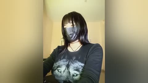 A video of a young Asian woman with long black hair, wearing a black mask and long-sleeved shirt featuring a graphic of wolf eyes, standing in a beige room with minimal decor.