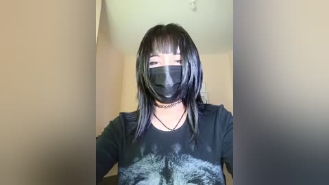 A video of an Asian woman with straight black hair, bangs, and a black face mask, wearing a black shirt with a white abstract design, a black choker, and a black chain necklace. The background is a beige wall and a white ceiling.