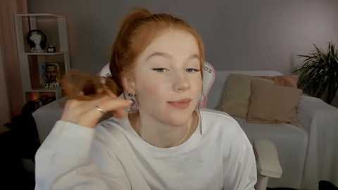 Media: Video of a young Caucasian woman with pale skin and red hair in a ponytail, wearing a white sweatshirt. She's seated in a cozy living room with a beige sofa, a green plant, and a bookshelf in the background.