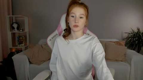 Video of a fair-skinned, red-haired woman in a white sweatshirt, wearing a pink headband, in a dimly lit living room with beige furniture and a bookshelf.