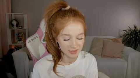 Media: Video of a young Caucasian girl with red hair in a ponytail, wearing headphones, sitting on a light gray couch in a cozy living room with beige walls, shelves, and a plant.