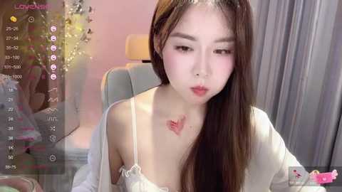 Media: A young Asian woman with fair skin, long brown hair, and light makeup sits in a modern room, wearing a white off-shoulder top, revealing a red dragon tattoo on her chest.