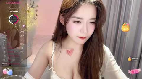 Media: Video of a young Asian woman with long brown hair, wearing a white lace camisole, in a soft pink room. She has a heart tattoo on her chest.