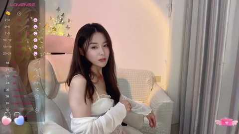 Media: A video of an East Asian woman with long black hair, fair skin, and delicate features, wearing a white lace dress, sitting on a bed in a softly lit room.