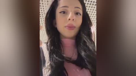 Video of a young woman with long black hair, light skin, and full lips, wearing a pink turtleneck sweater, against a beige and white tiled background.