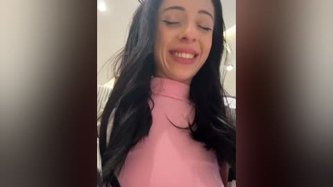 Video of a smiling woman with long black hair and fair skin, wearing a pink turtleneck sweater, against a blurred, neutral-toned background.