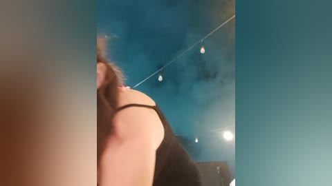 Media: Video of a woman with brown hair, wearing a black tank top, seen from behind, in a dimly lit room with string lights and a blue wall.