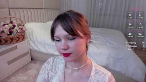 Media: Video of a fair-skinned woman with brown hair and red lipstick, wearing a white lace dress and pearl necklace, sitting on a plush carpet in a modern bedroom with a white bed and floral basket.