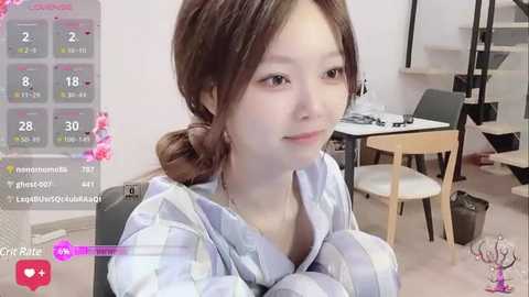 Media: Video of a young Asian woman with light skin and brown hair in a ponytail, wearing a striped robe, sitting in a modern, minimalist room with a dining table and chairs.