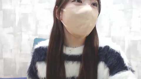 Media: Video of an East Asian woman with long black hair, wearing a black and white striped sweater and a face mask, in a blurred, softly lit room with white curtains.