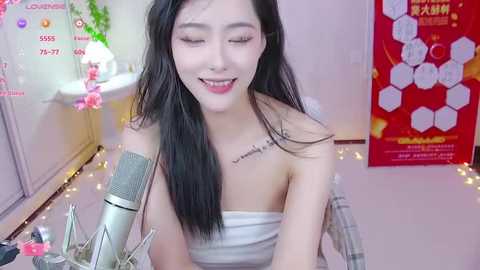 Media: Video of a smiling East Asian woman with long black hair, wearing a white off-shoulder top, sitting at a pink-tiled desk with a microphone, surrounded by pink cherry blossom decor.