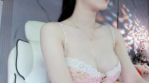 Media: Video of a pale-skinned woman with long black hair, wearing a lacy pink bra and necklace, seated on a white chair, with a blurred background of a marble wall and a Buddha statue.