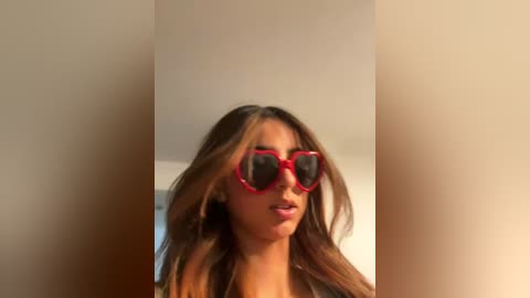 Media: Video of a young woman with long, wavy brown hair wearing red heart-shaped sunglasses indoors against a beige wall. She has a light skin tone and is looking slightly to the left.