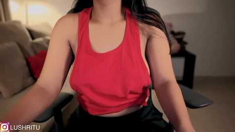 Media: Video of a woman with long dark hair wearing a tight red tank top, sitting on a chair in a dimly lit living room.