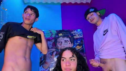 Media: Video of three young people in a colorful room. Two shirtless men lift their shirts, revealing their bodies. One woman in a Nike sweatshirt smiles in the foreground.