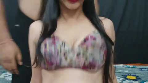 Media: Video of a young Asian woman with long black hair, wearing a colorful floral bra, indoors against a dark curtain backdrop. Her lips are painted bright red.