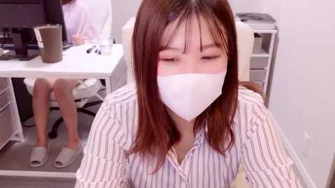 Media: Video of an Asian woman with straight, shoulder-length brown hair, wearing a white face mask, striped shirt, and slippers, sitting at a desk in a modern office with a computer monitor and white chair.