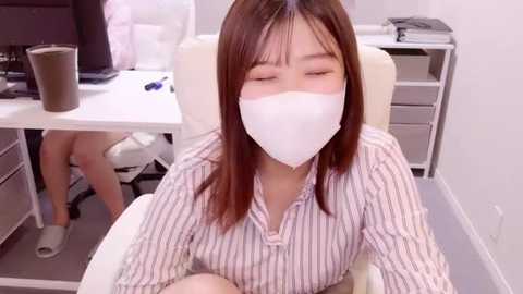 Media: Video of an Asian woman with straight, shoulder-length brown hair, wearing a white face mask, sitting in an office chair. She has a light complexion and is wearing a white and black striped blouse. The office background includes a desk with a computer monitor and a brown coffee cup.