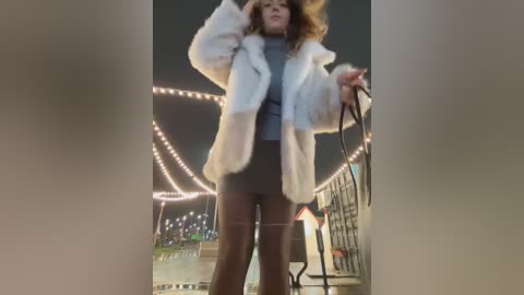 Media: Video of a woman with curly hair in a white fur coat, black leggings, and a gray top, standing in a night-time urban setting with string lights and a metal fence in the background.