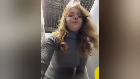 Media: A video of a young woman with long, wavy blonde hair and fair skin, wearing a grey turtleneck sweater, standing in a dimly lit elevator with a metallic, grid-patterned ceiling.