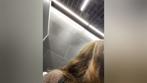 Media: A video showing a close-up of a person's shoulder and back, facing away, with long, wavy blonde hair. The background features a modern, industrial-style interior with gray metallic panels and fluorescent lighting.