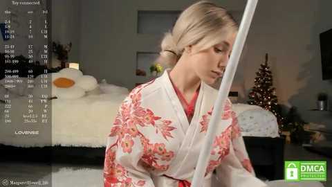 Media: Video of a blonde woman in a floral kimono, looking down, in a cozy, Christmas-decorated bedroom with a TV screen displaying a calendar.