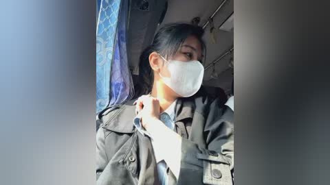 Media: Video of an Asian woman with long black hair, wearing a white face mask and black jacket, seated on a bus. The background features blue and gray seat covers and overhead luggage racks.