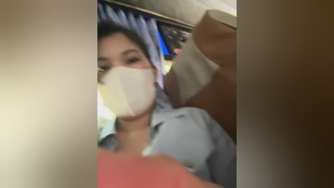 Media: A video of a young Asian woman with dark hair, wearing a light blue shirt and a white face mask, taking a selfie inside a dimly lit room with a blue curtain in the background.